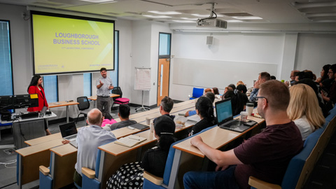 People seated in a tiered classroom, facing a lecturer with a projector screen displaying 